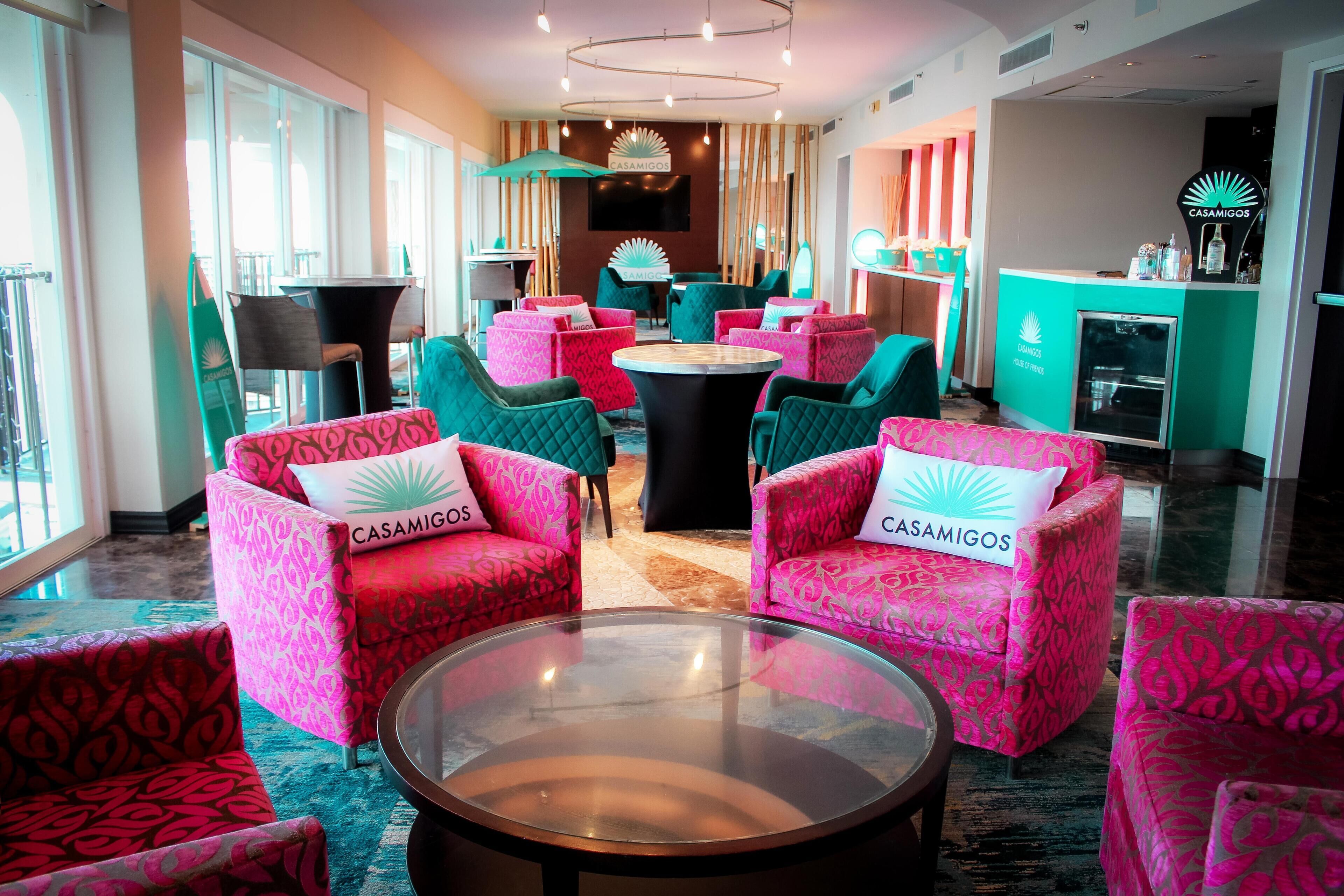 Un hall d’hôtel avec des fauteuils à motifs turquoise et rose, une table ronde centrale et un comptoir de réception doté d’un téléviseur et d’une enseigne Casamigos.