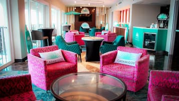 Eine Hotellobby mit sesseln in türkisfarbenem und rosaem Muster, einem runden Tisch in der Mitte und einer Rezeption mit Fernseher und Casamigos-Beschilderung.