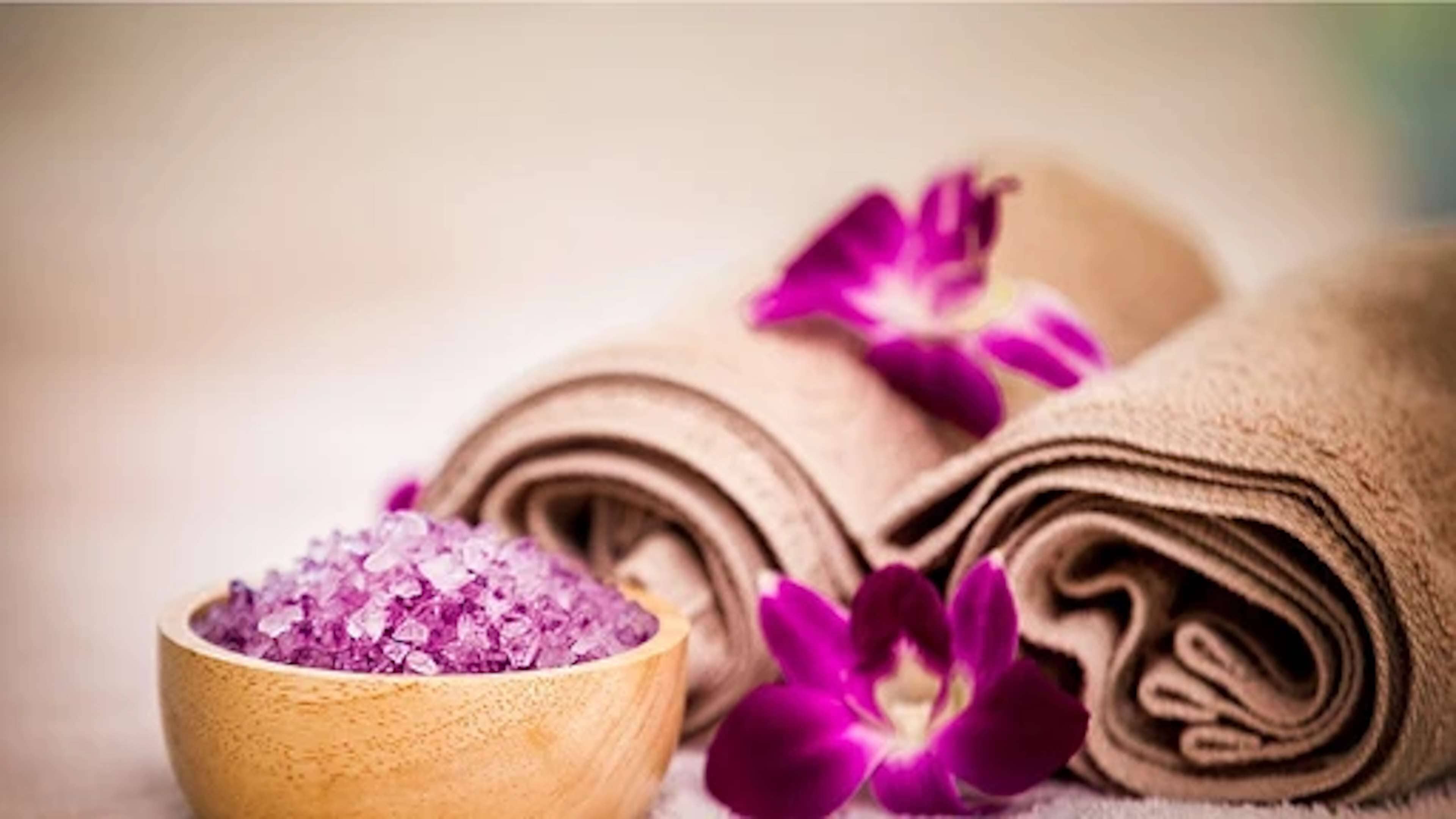 A wooden bowl filled with purple bath salts, rolled towels, and purple orchid flowers.