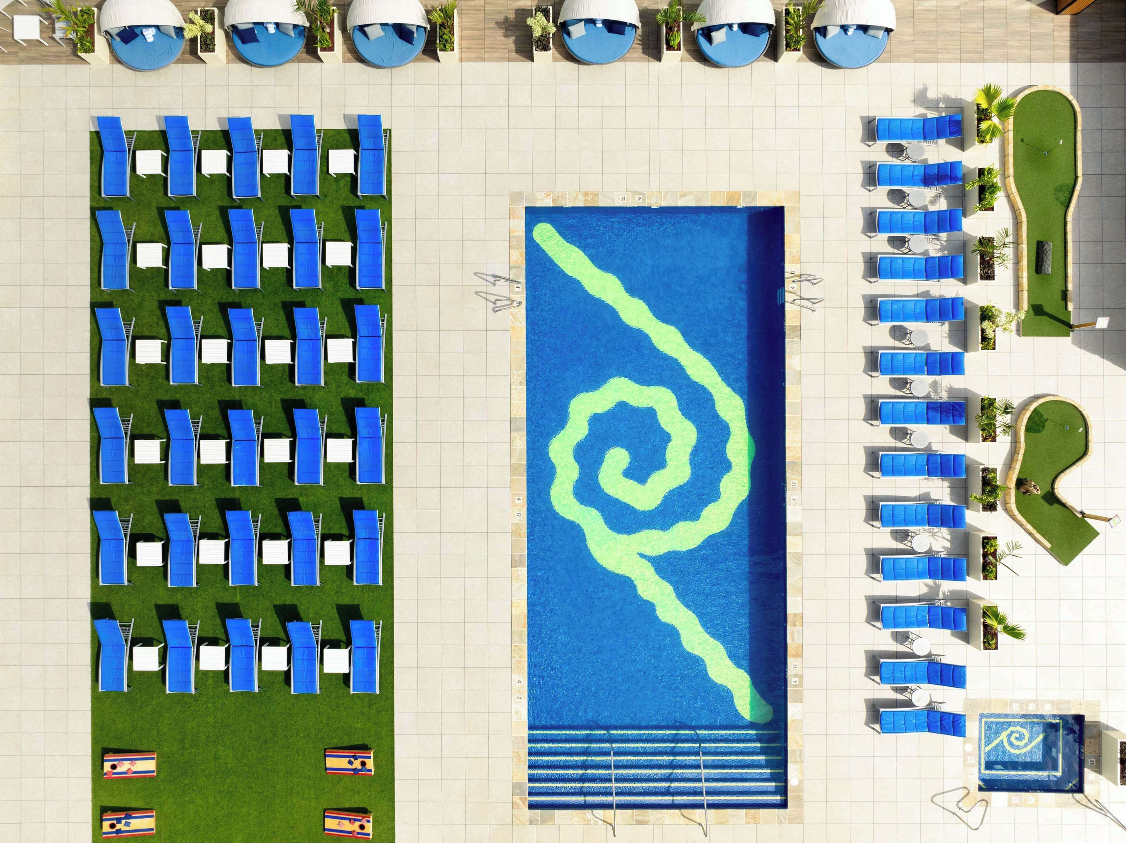 An aerial view of a hotel pool area with multiple lounge chairs and umbrellas, a central swimming pool with a distinctive design, and a small putting green.