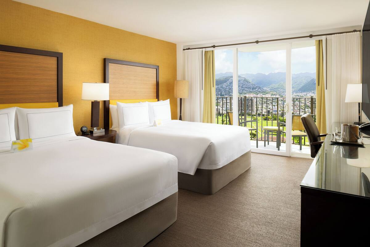 A hotel room with two beds, a desk, and a balcony with a city view.