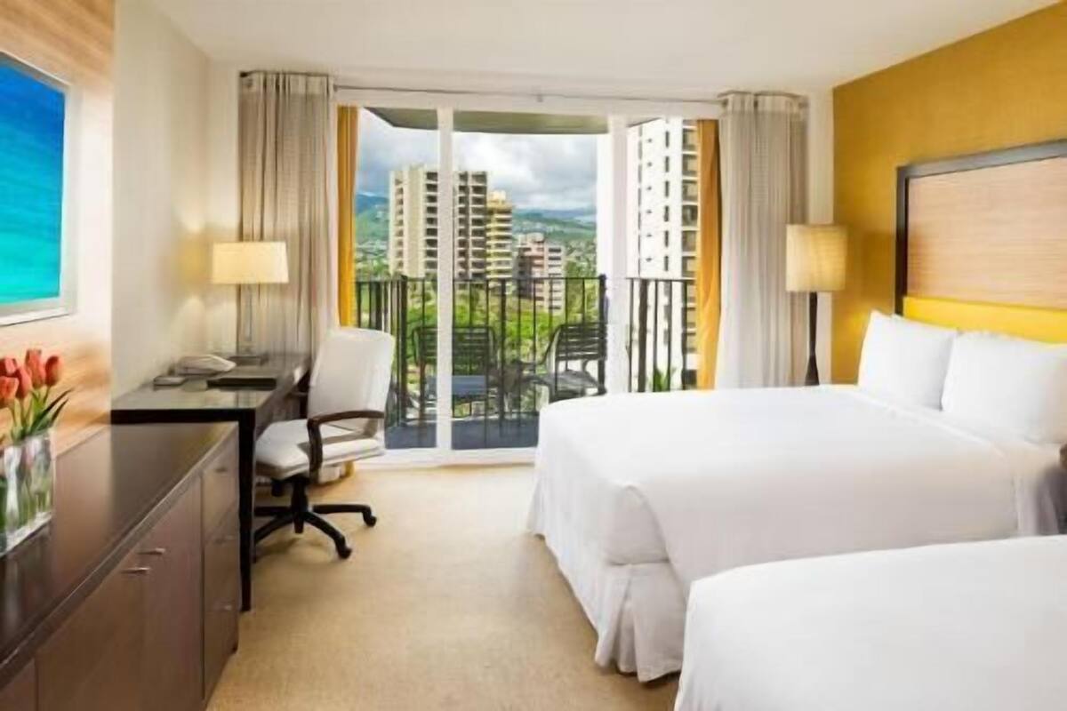 A hotel room with a bed, a desk, a chair, and a view of buildings through a balcony.