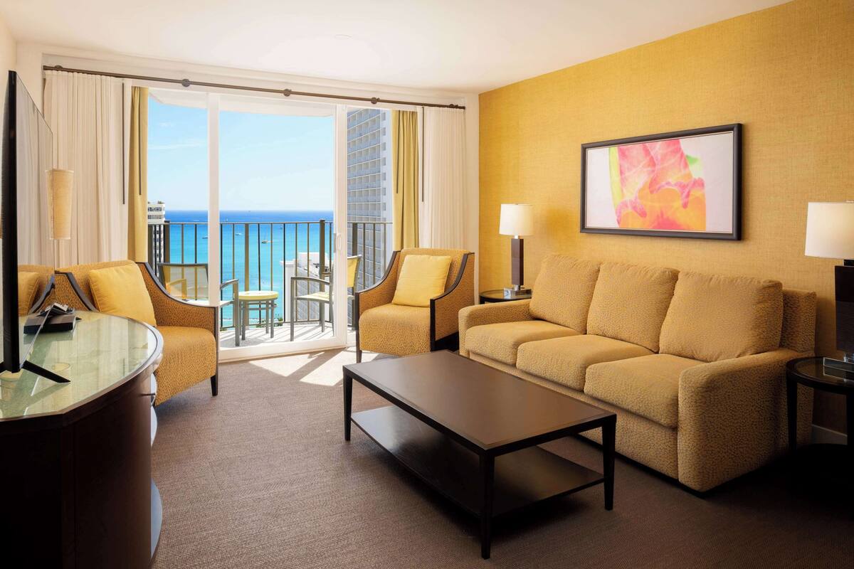 A hotel room with a sofa, armchairs, a coffee table, a glass table, and a balcony with a view of the sea.