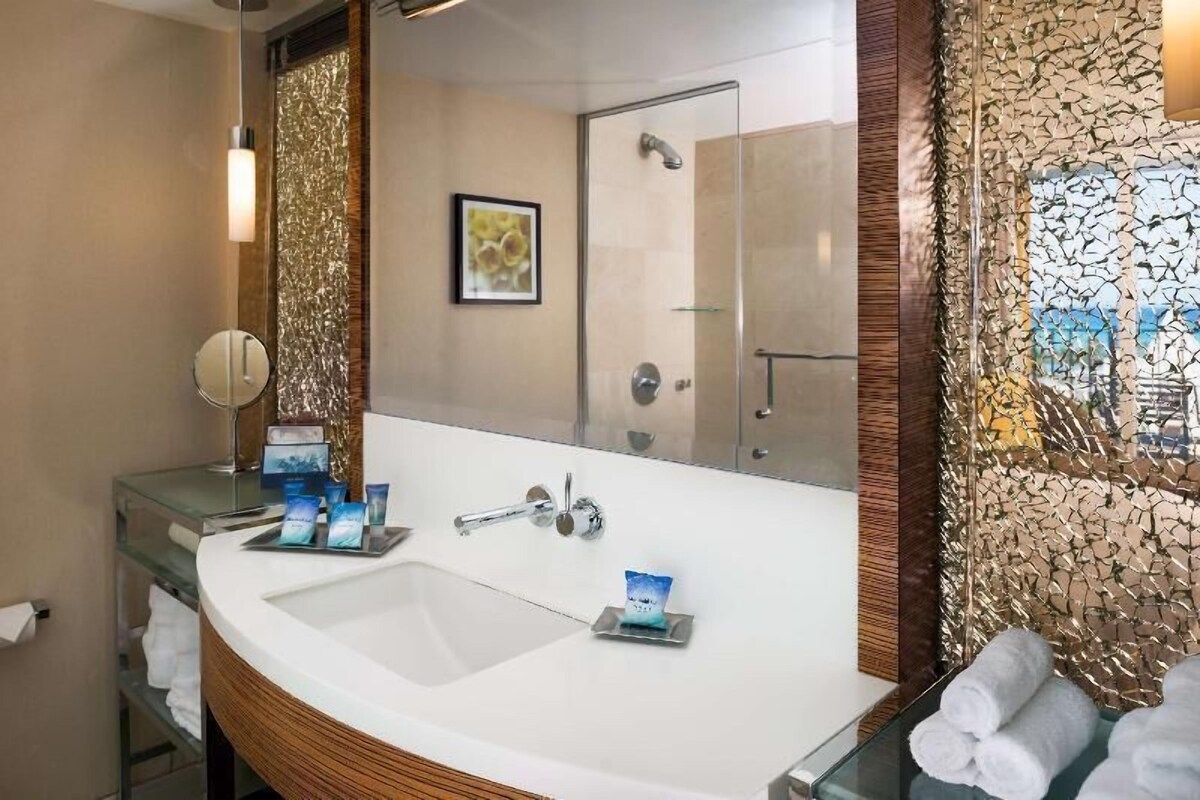 A bathroom with a large mirror, a sink with toiletries, a shower, and a decorative wall.