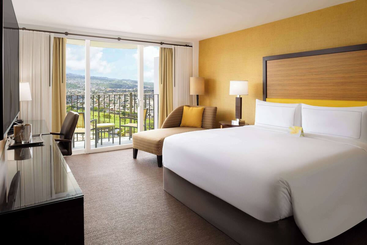 A hotel room with a large bed, a desk, a chair, and a view of a cityscape through large windows.