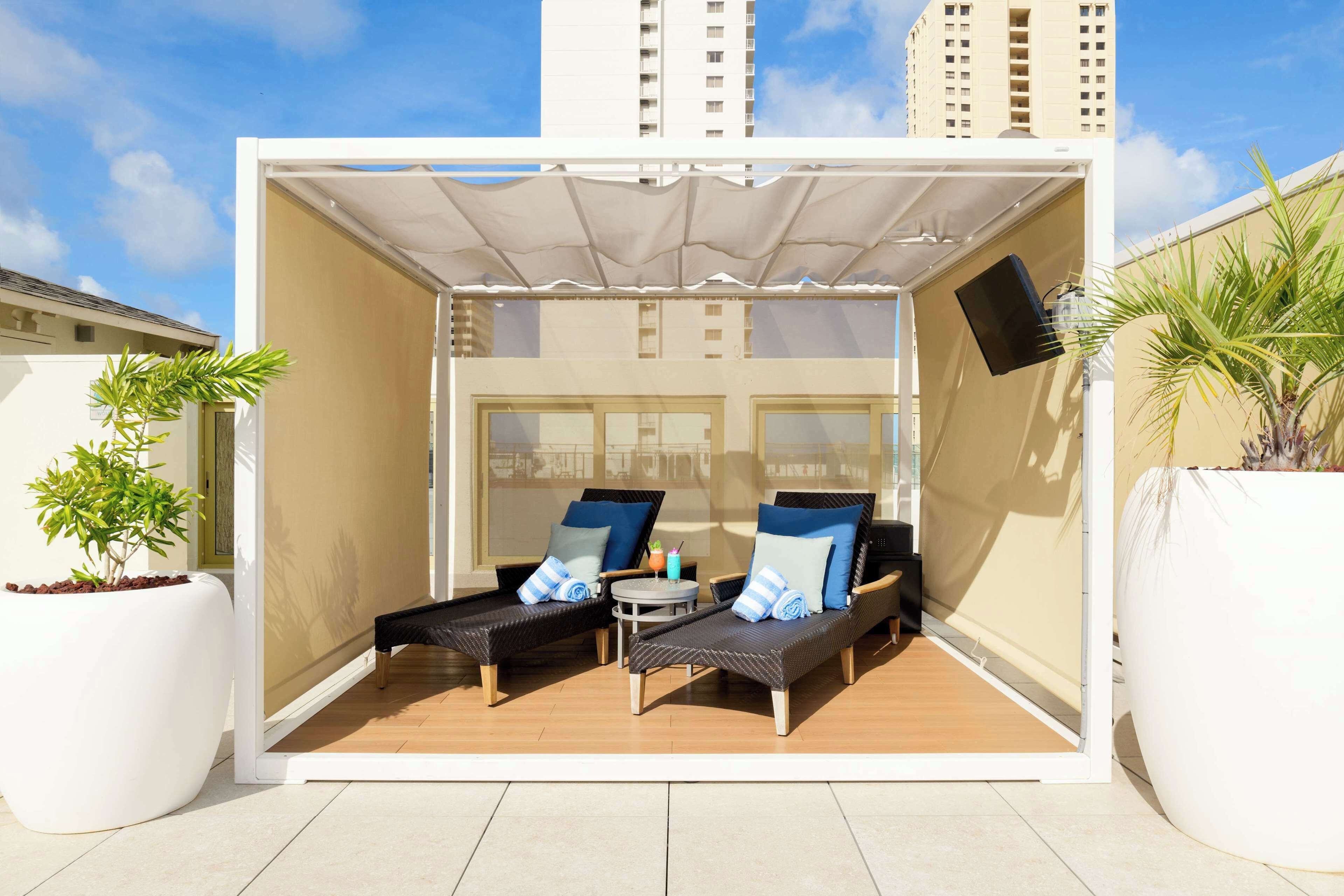 A small outdoor lounge area with two black lounge chairs, a small table, and a white umbrella providing shade.