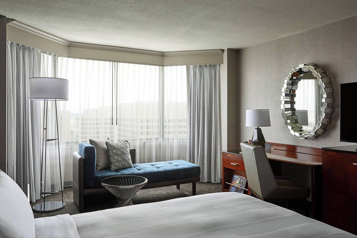 A hotel room with a bed, a desk, a chair, a small sofa, a lamp, a mirror, and large windows with curtains.