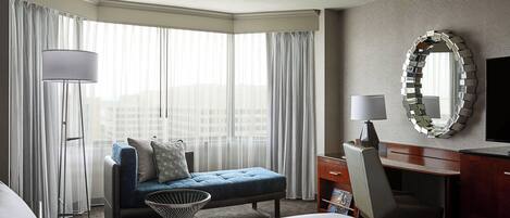 Habitación de hotel con cama, escritorio, silla, un pequeño sofá, lámpara, espejo y ventanas amplias con cortinas.