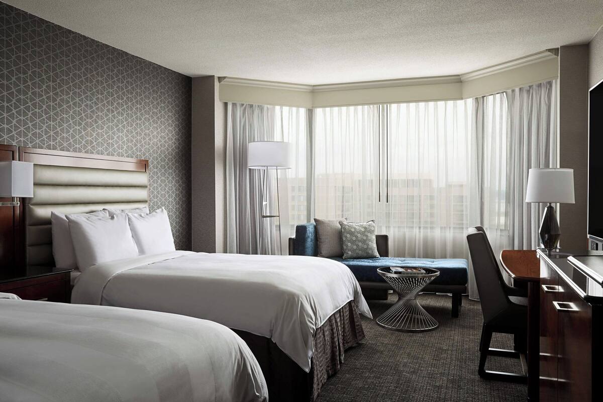 A hotel room with two beds, a desk, a chair, and a small table.