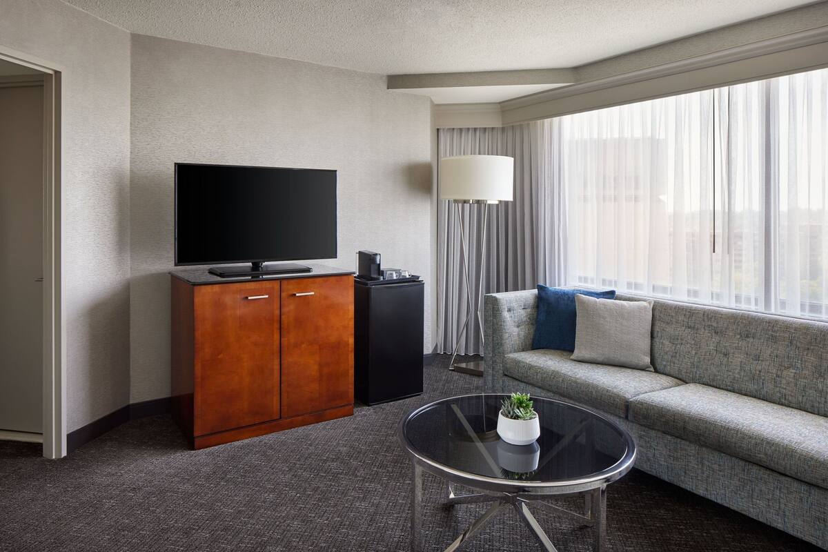 A modern hotel room with a flat-screen TV, a wooden cabinet, a sofa, a glass coffee table, and a large window with sheer curtains.