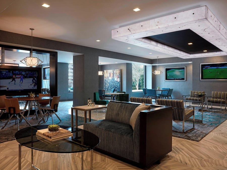 A spacious lobby with a modern design, featuring a seating area with sofas and chairs, a dining table, and large screens displaying sports.