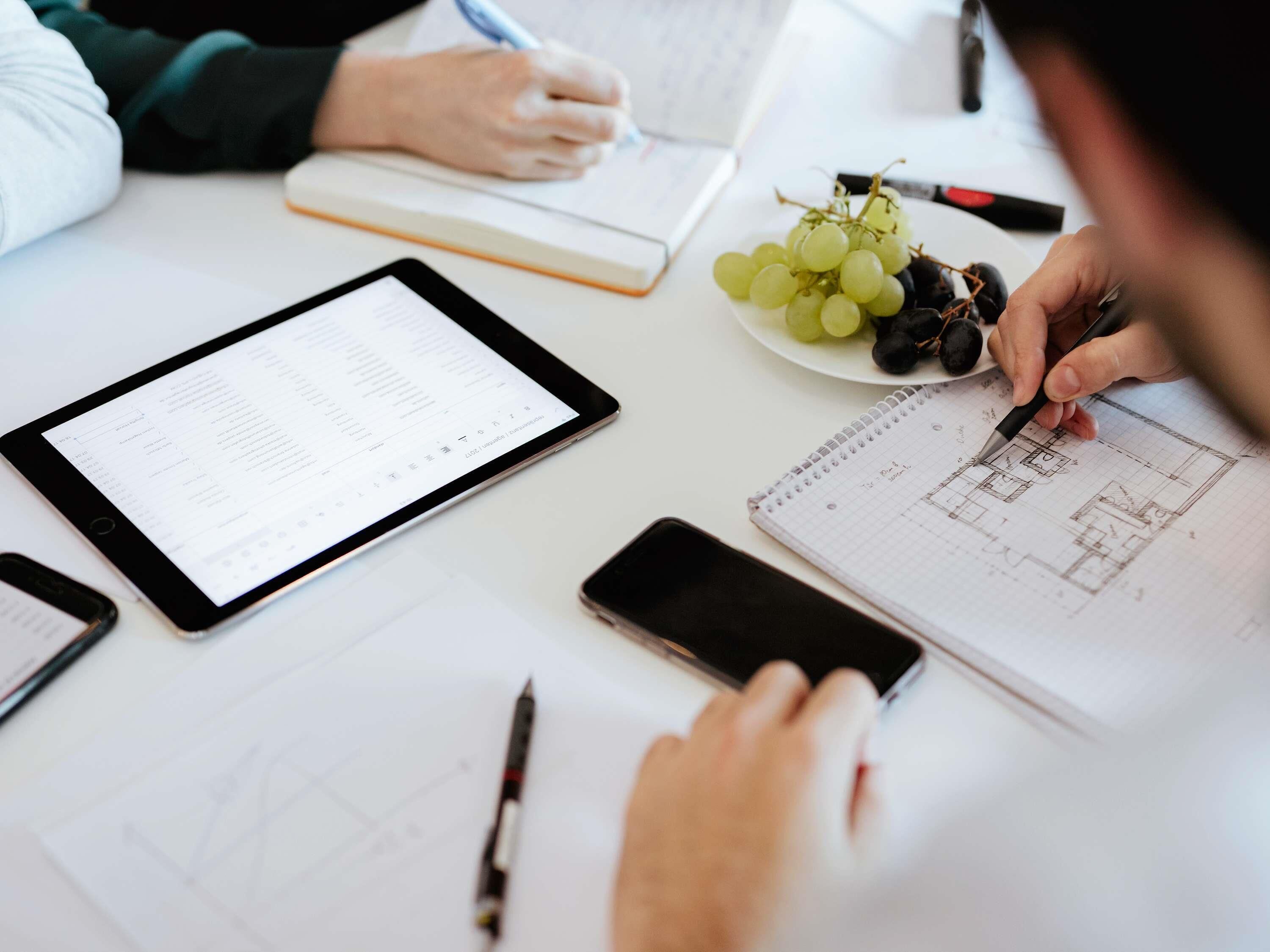 A person writing on a notebook with a pen, a tablet displaying a spreadsheet, and a plate of grapes on a table.