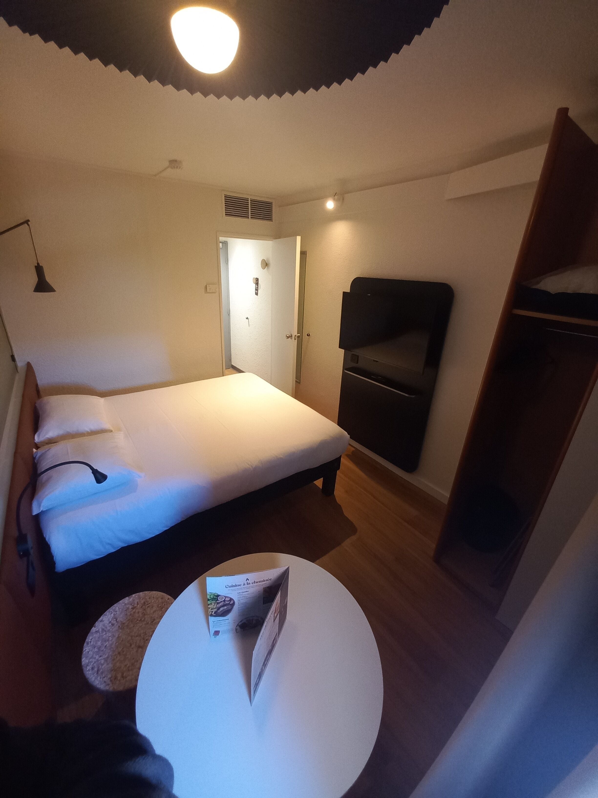 A hotel room with a bed, a small round table, a TV, a wardrobe, and a door leading to another room.