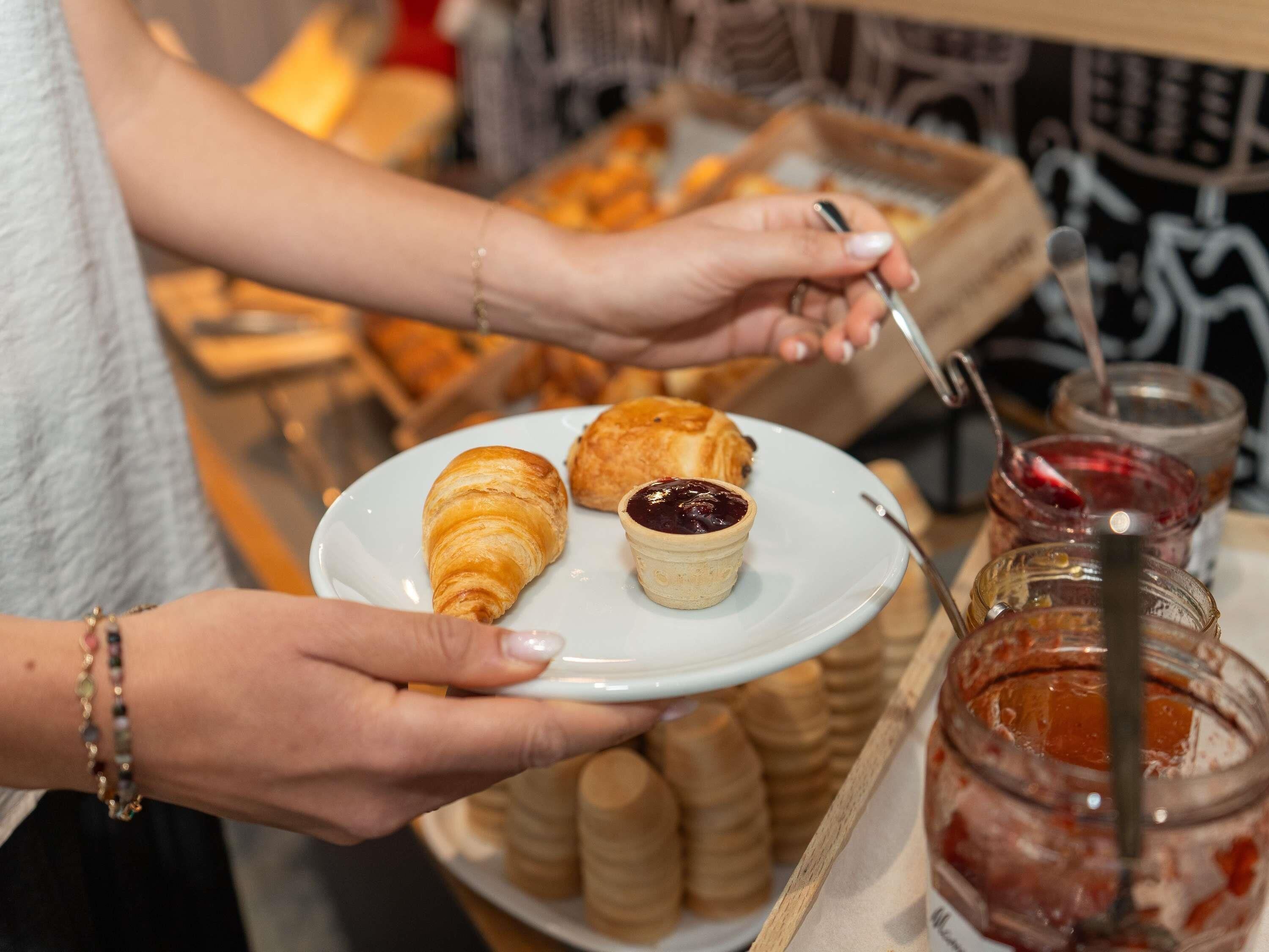 Una persona sosteniendo un plato con un croissant y un recipiente pequeño de mermelada, con un frasco de mermelada al fondo.
