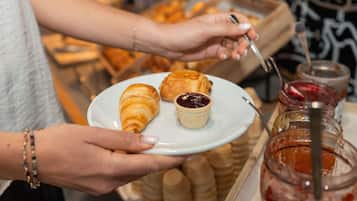 Una persona sosteniendo un plato con un croissant y un recipiente pequeño de mermelada, con un frasco de mermelada al fondo.