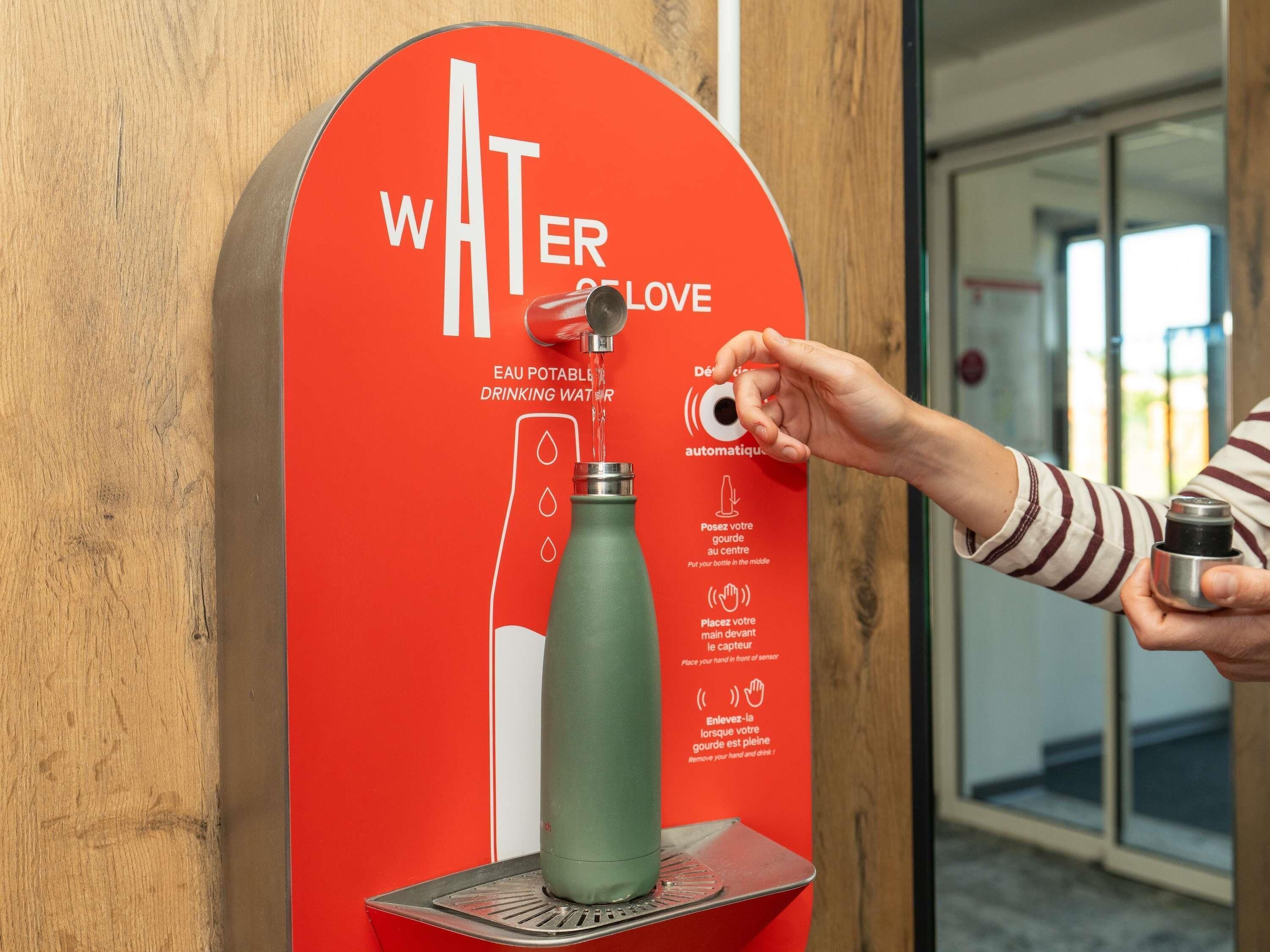 A water dispenser with instructions in multiple languages, a hand holding a bottle, and a red sign with white text.