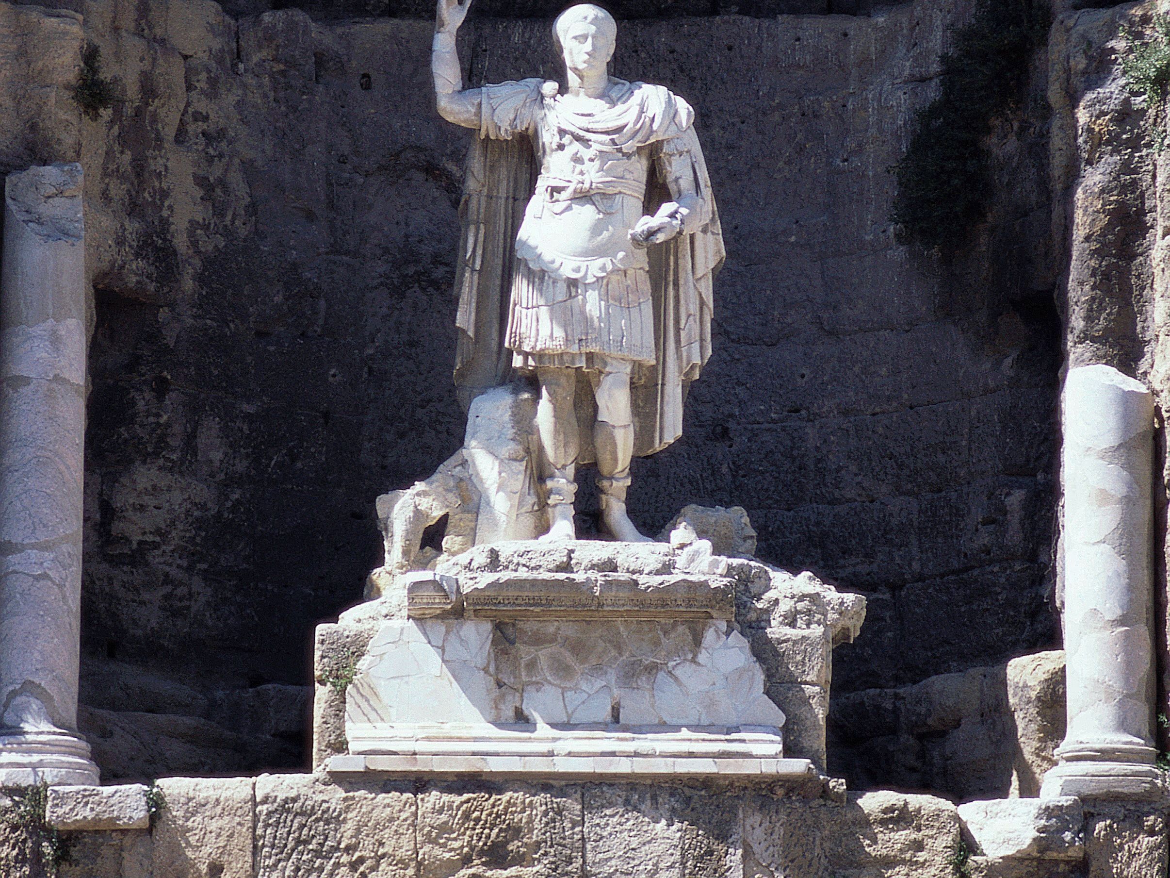 A marble statue of a historical figure on a pedestal with a stone base, surrounded by ancient stone structures.