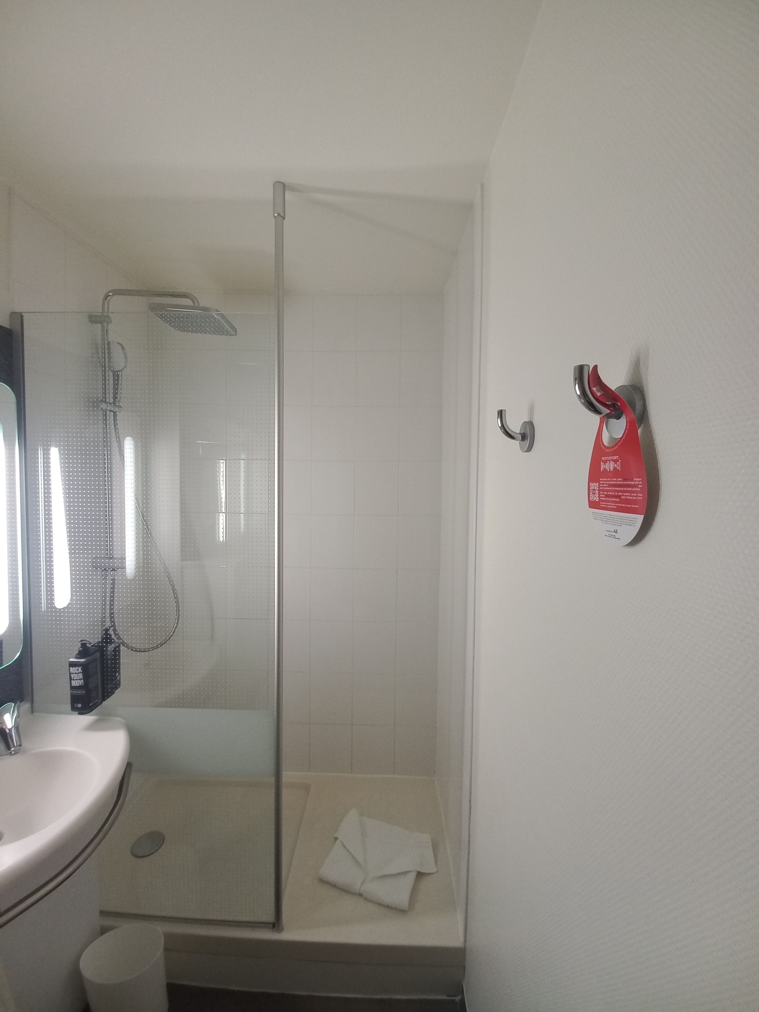 A bathroom with a glass shower enclosure, a sink, a towel, and a red safety hook on the wall.
