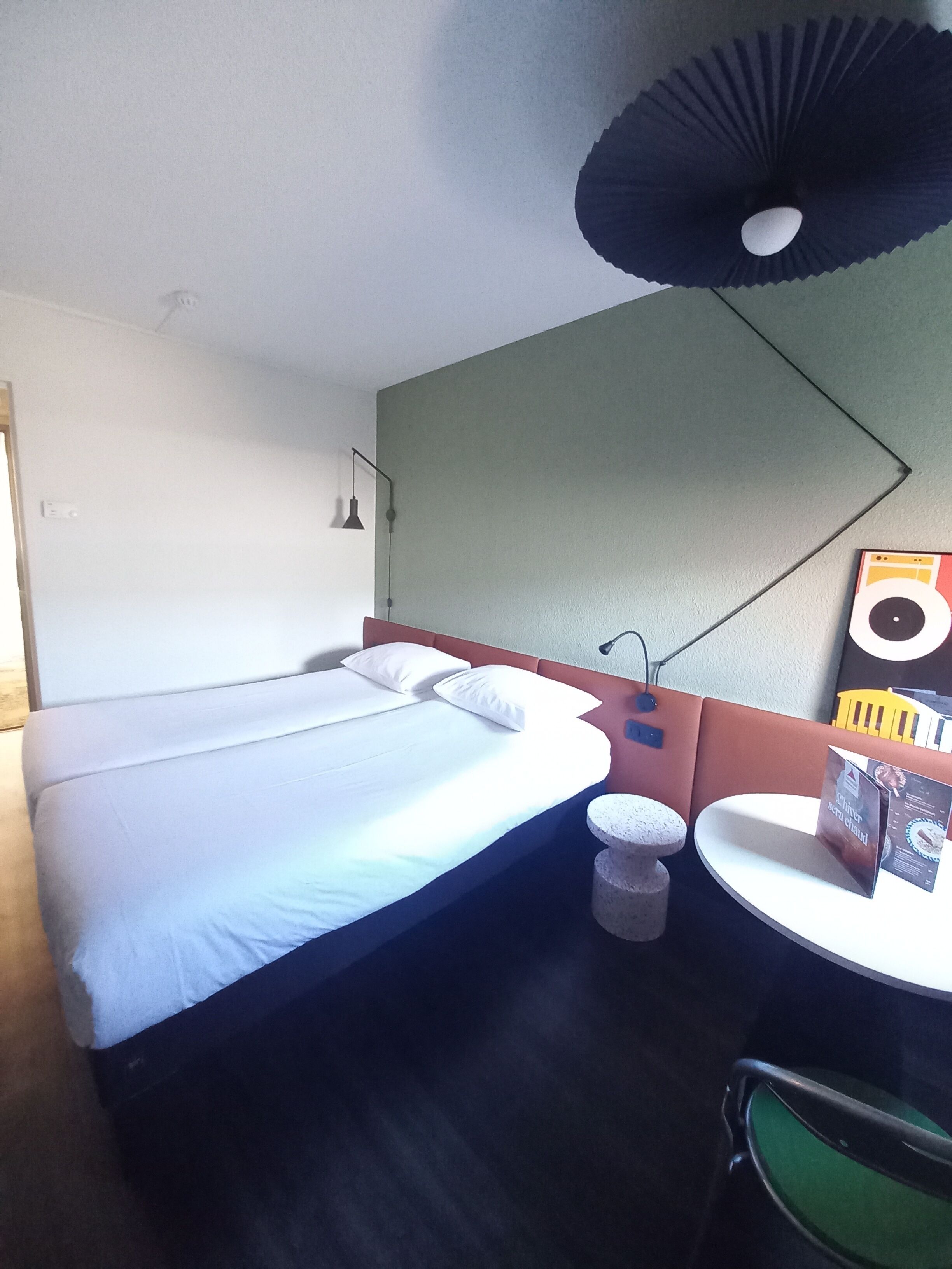 A hotel room with a bed, a small round table, a chair, a lamp, and a wall-mounted mirror.