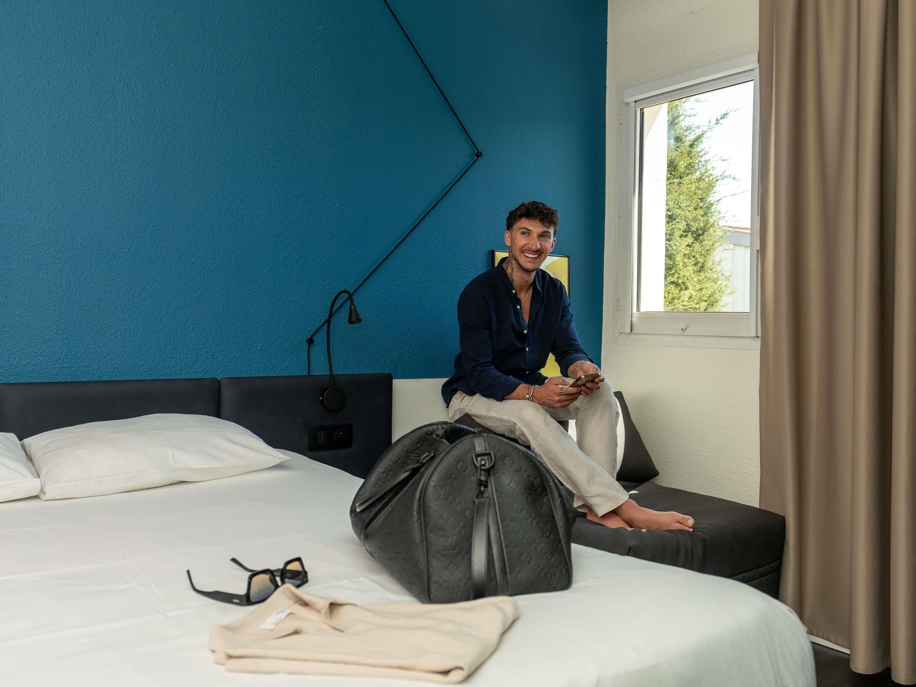Une chambre d’hôtel avec un lit, une fenêtre et une personne assise sur le lit.