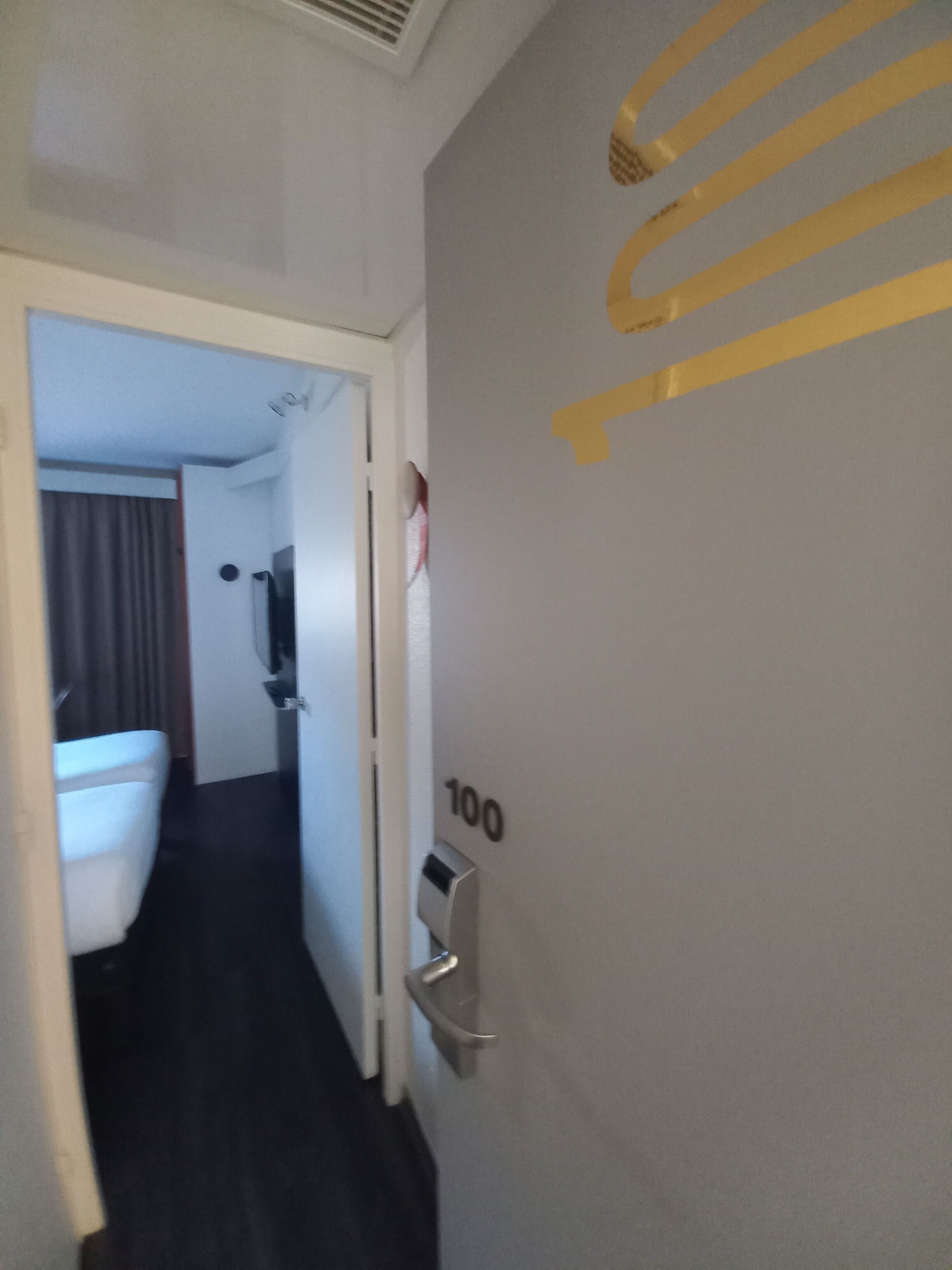 Een hotelgang met een deur met het nummer 100 en een zichtbare badkamer met toilet en spiegel.