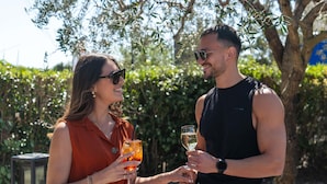 Dos personas brindando con bebidas en un entorno exterior con árboles y un muro de piedra.