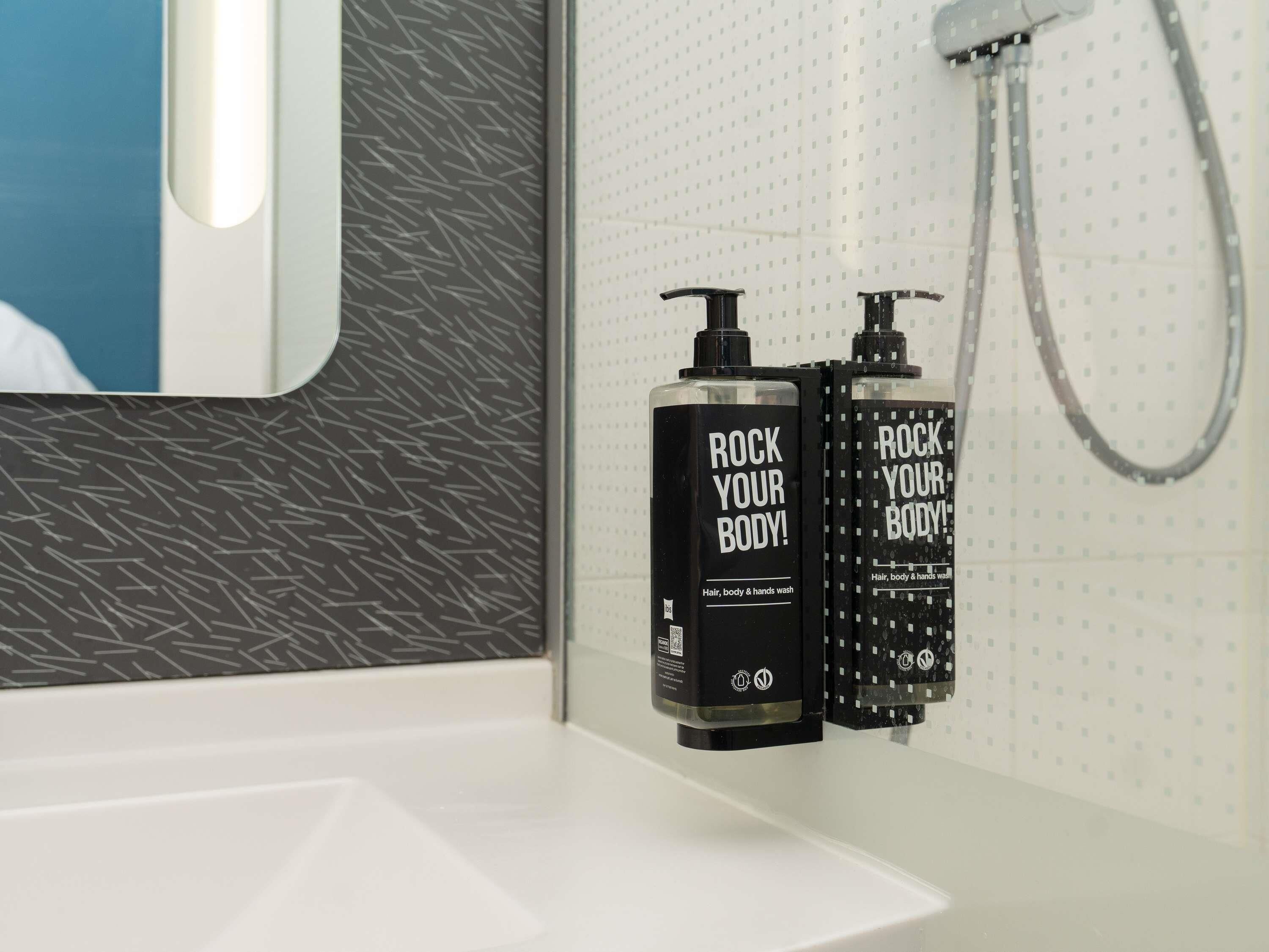 Een badkamer met een witte wastafel, twee zwarte flesjes met de tekst 'ROCK YOUR BODY!' voor haar-, lichaams- en handverzorging, en een douchekop.
