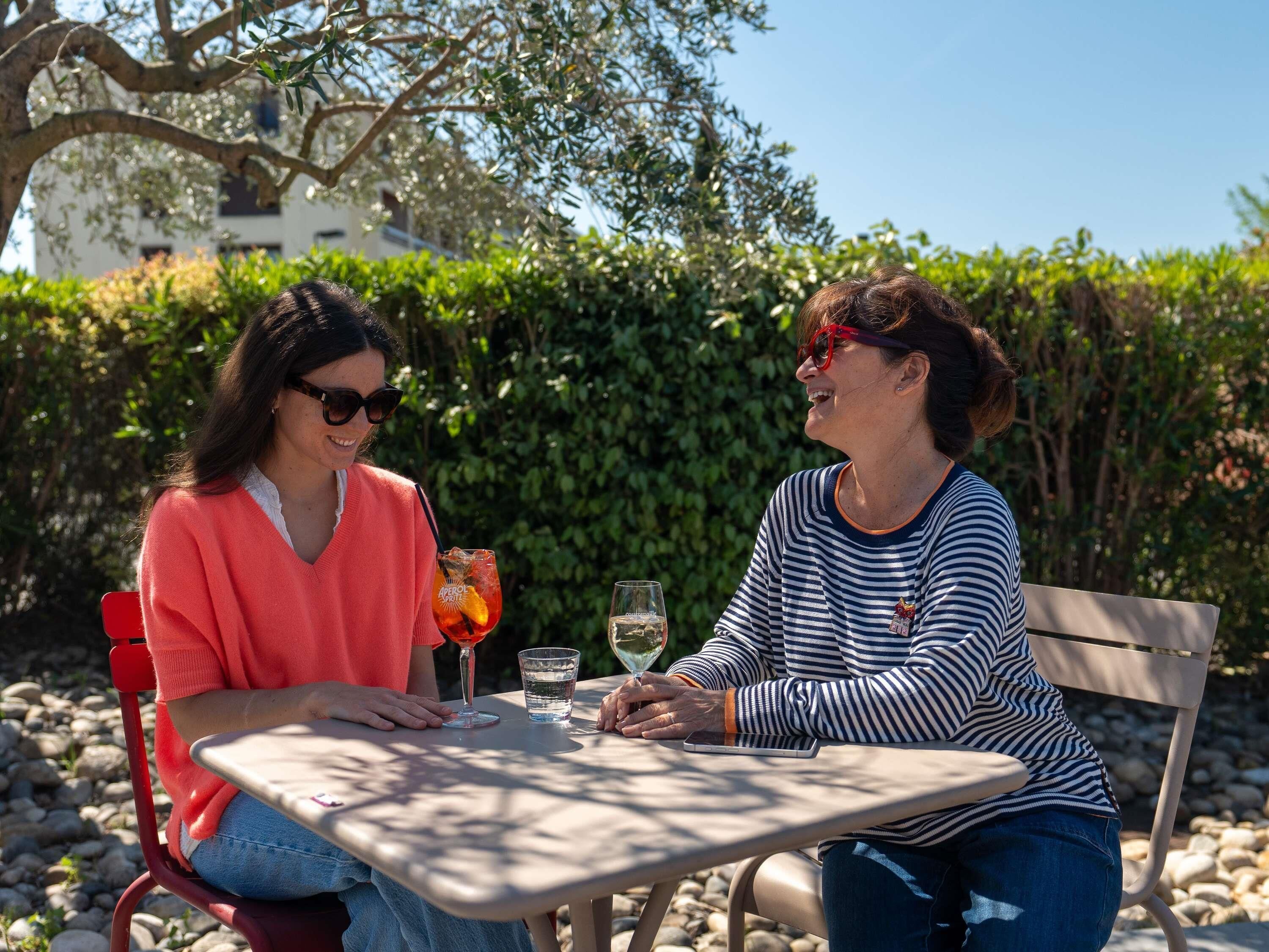 Two people seated at an outdoor table with drinks, surrounded by greenery and a building in the background.
