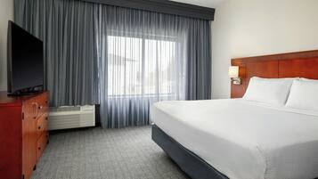 Ein Hotelzimmer mit einem großen Bett, einem Fernseher auf einer Holzkommode, einem Fenster mit Vorhängen und einer Wandleuchte.