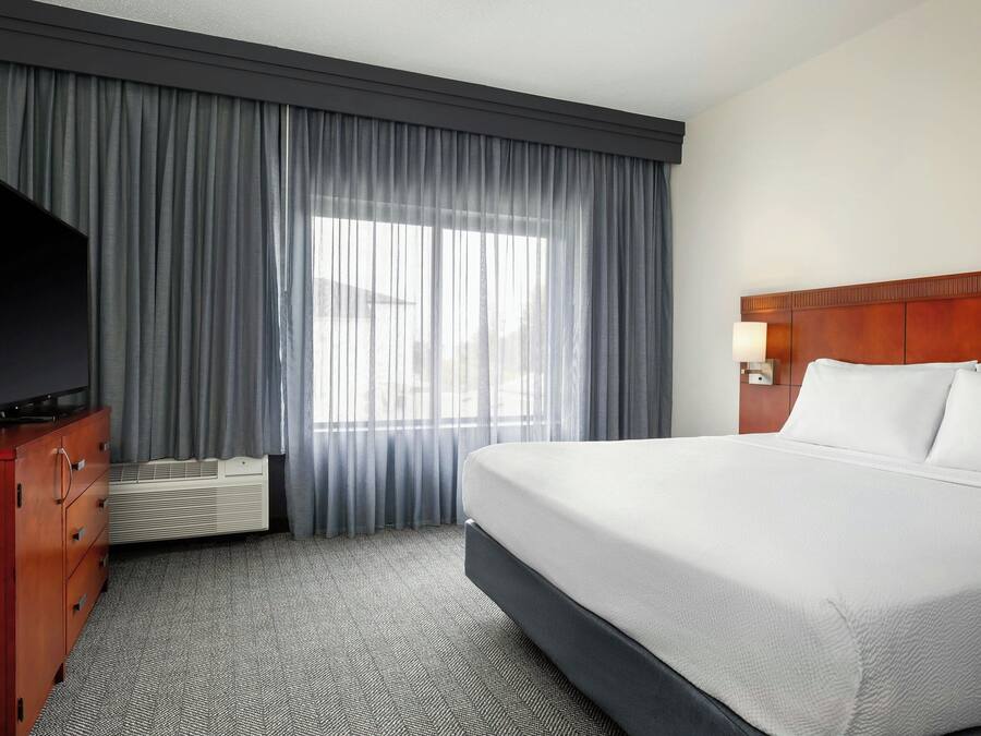 Ein Hotelzimmer mit einem großen Bett, einem Fernseher auf einer Holzkommode, einem Fenster mit Vorhängen und einer Wandleuchte.