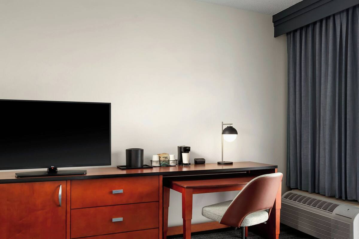 A hotel room with a flat-screen TV, a desk with a chair, a coffee maker, a lamp, and a window with curtains.