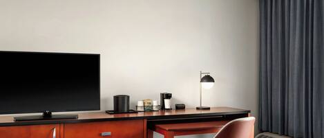 A hotel room with a flat-screen TV, a desk with a chair, a coffee maker, a lamp, and a window with curtains.