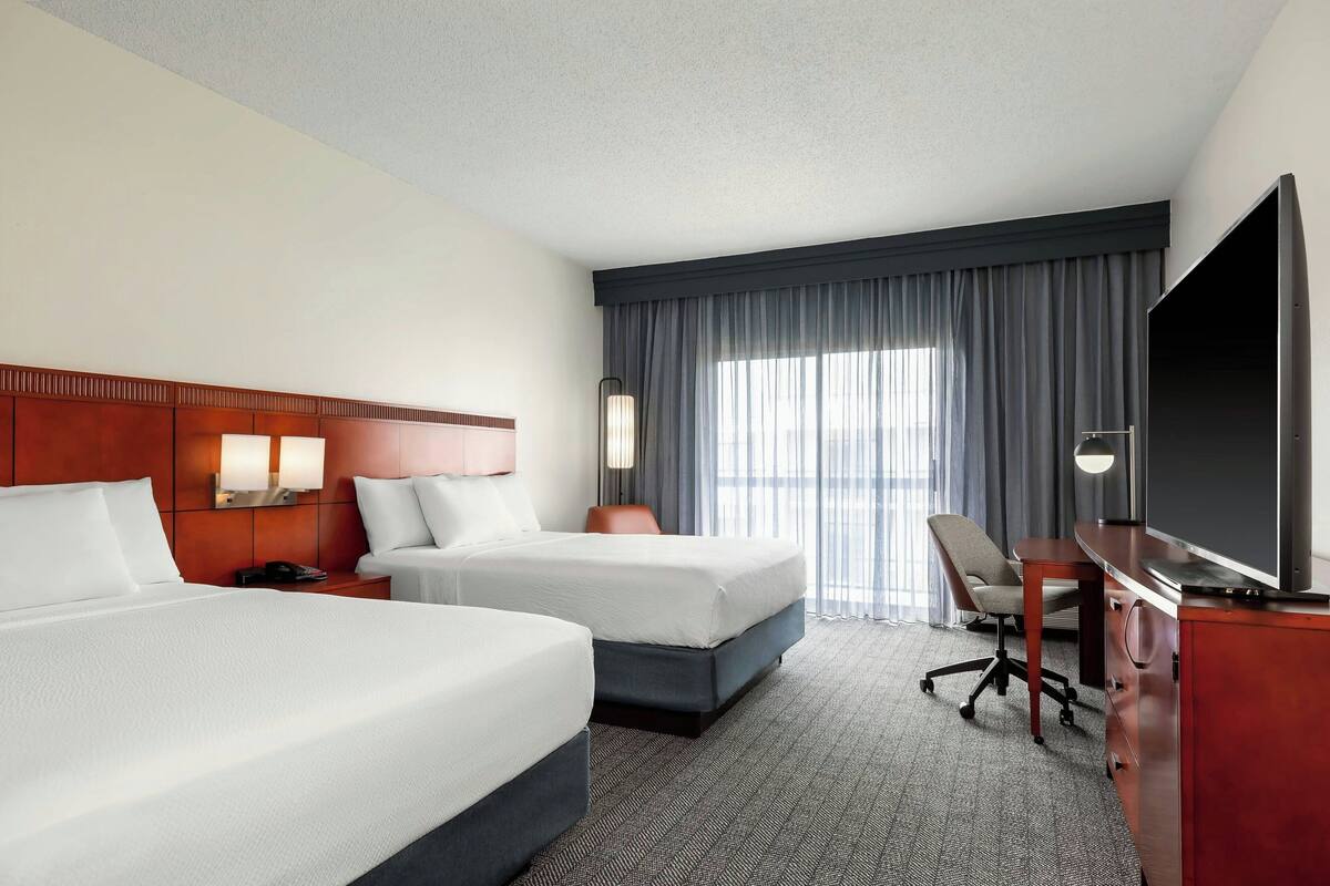 A hotel room with two beds, a desk, a TV, and a large window with curtains.