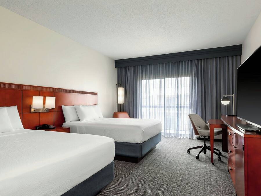 A hotel room with two beds, a desk, a TV, and a large window with curtains.