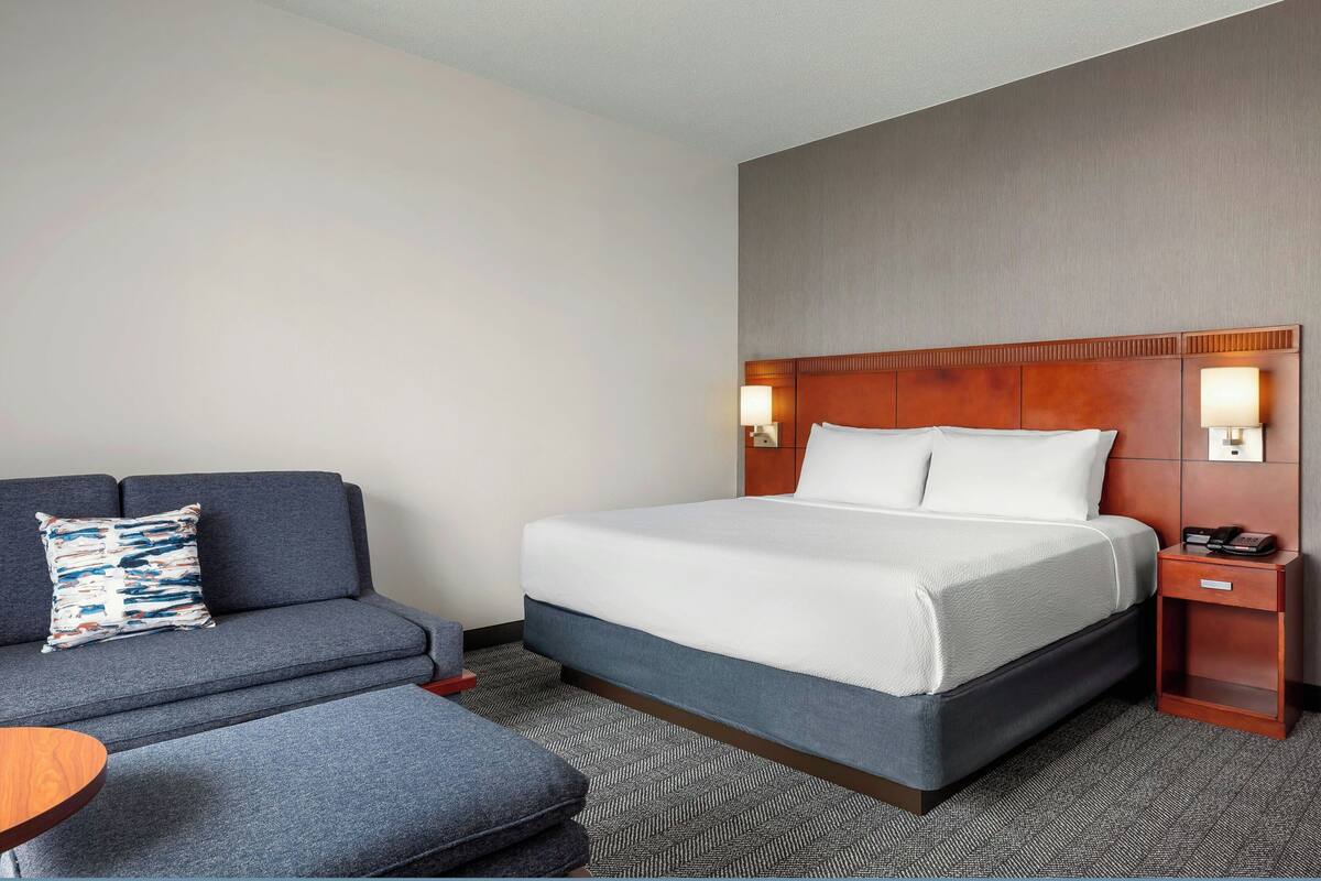 A hotel room with a bed, a sofa, a small table, and a nightstand with a lamp.