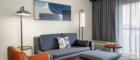 Una habitación de hotel moderna con un sofá gris, un sillón naranja, una mesita de madera y una lámpara de pie.