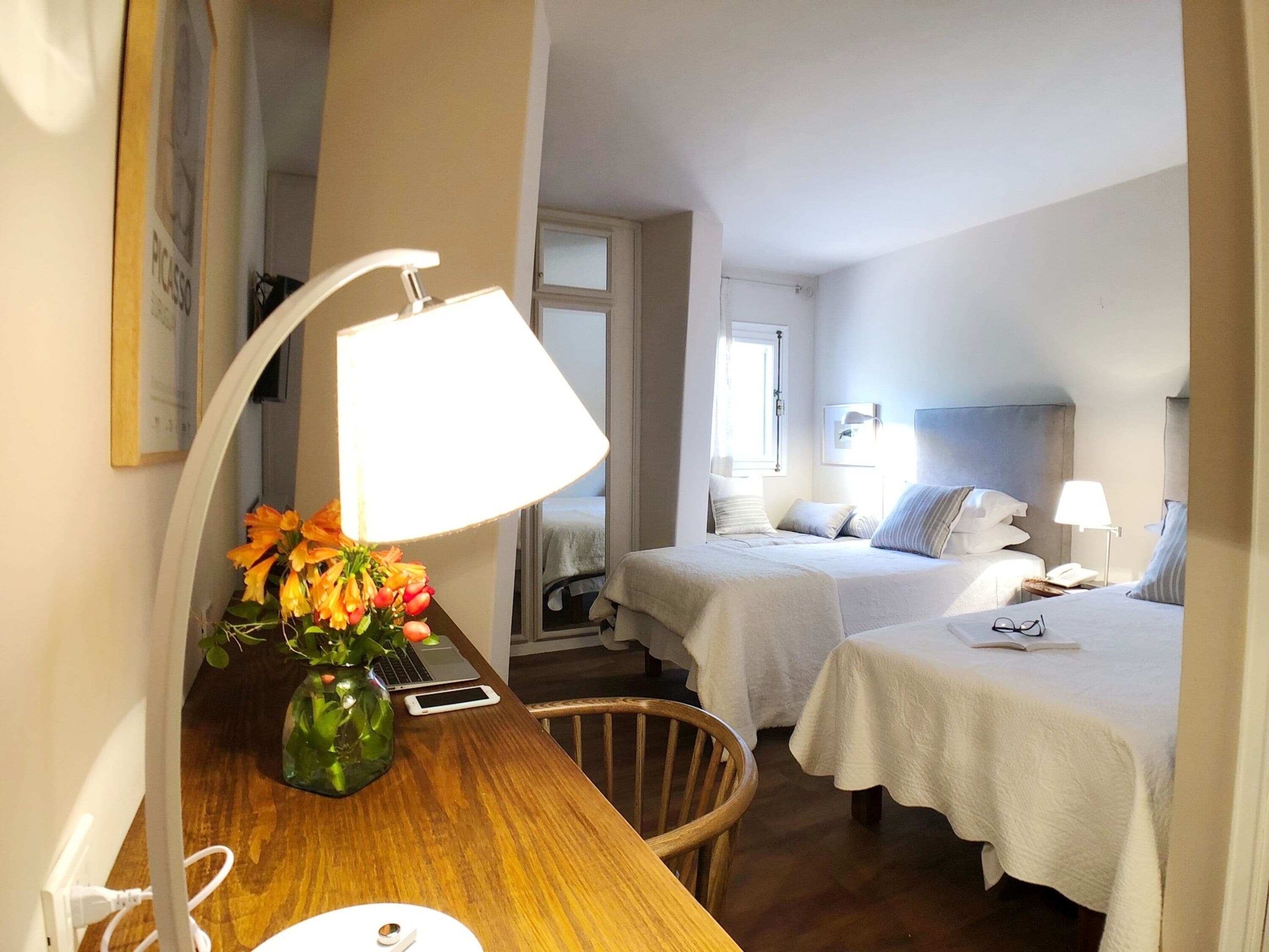 A hotel room with two beds, a desk with a lamp, a chair, and a vase with flowers.