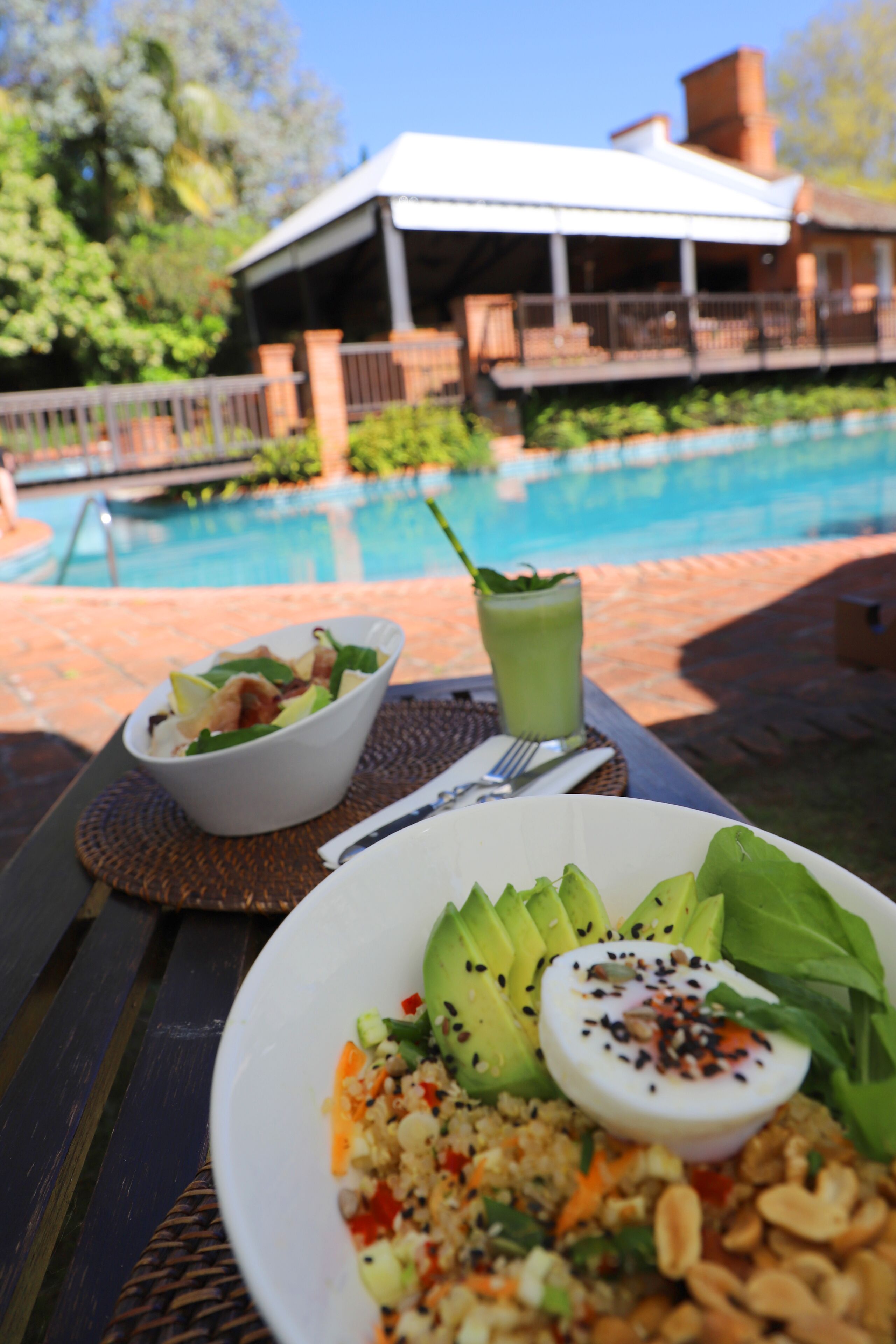 Una mesa de comedor con una ensalada, una bebida verde y un plato de comida. Al fondo, se ve una piscina y un edificio con porche cubierto.