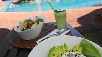 Una mesa de comedor con una ensalada, una bebida verde y un plato de comida. Al fondo, se ve una piscina y un edificio con porche cubierto.