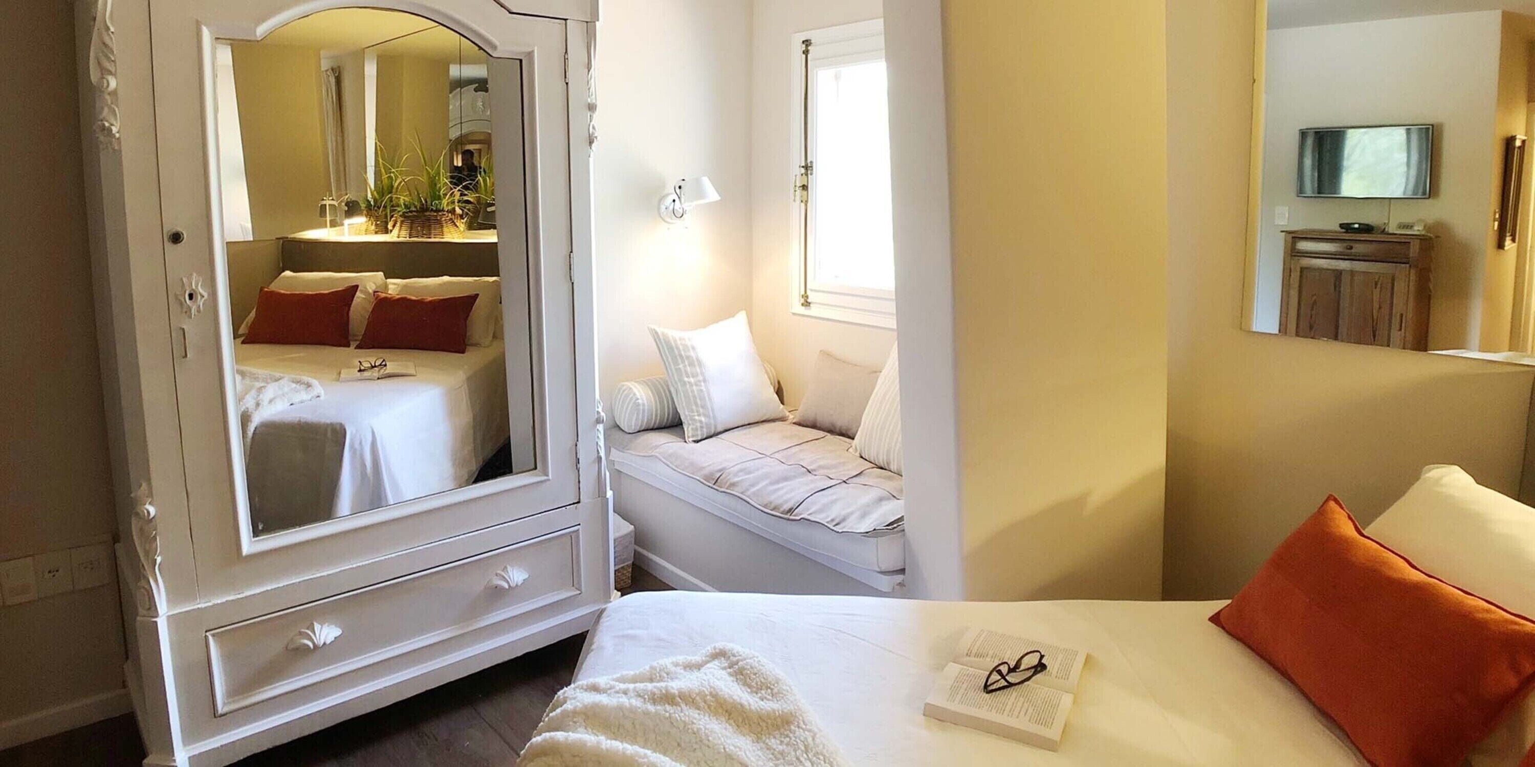Une chambre avec un grand miroir, une table de chevet sur laquelle se trouve un livre, une fenêtre avec des rideaux et un téléviseur posé sur une commode en bois.