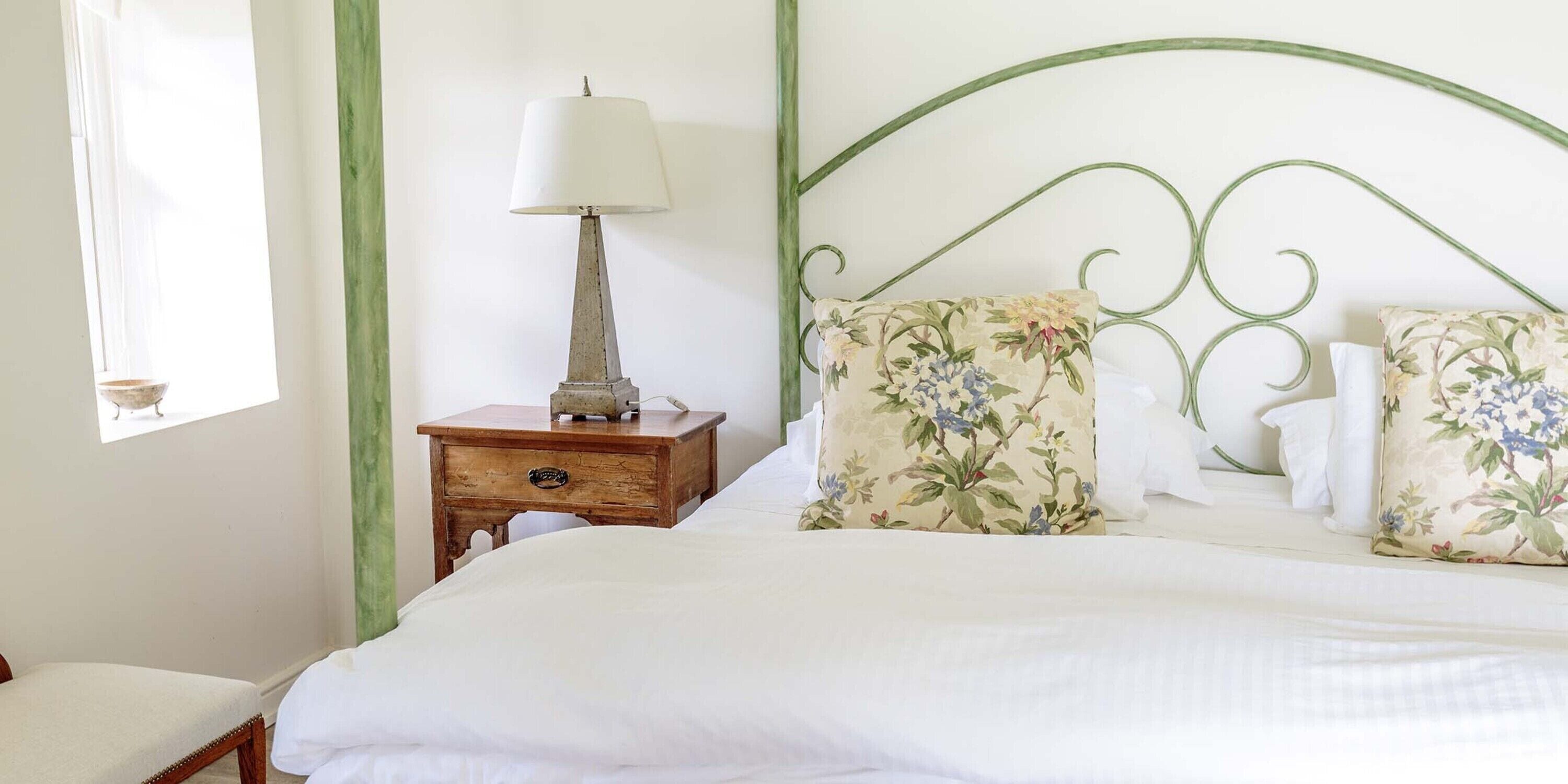 A neatly made bed with floral pillows, a bedside table with a lamp, and a window with a view of the outdoors.