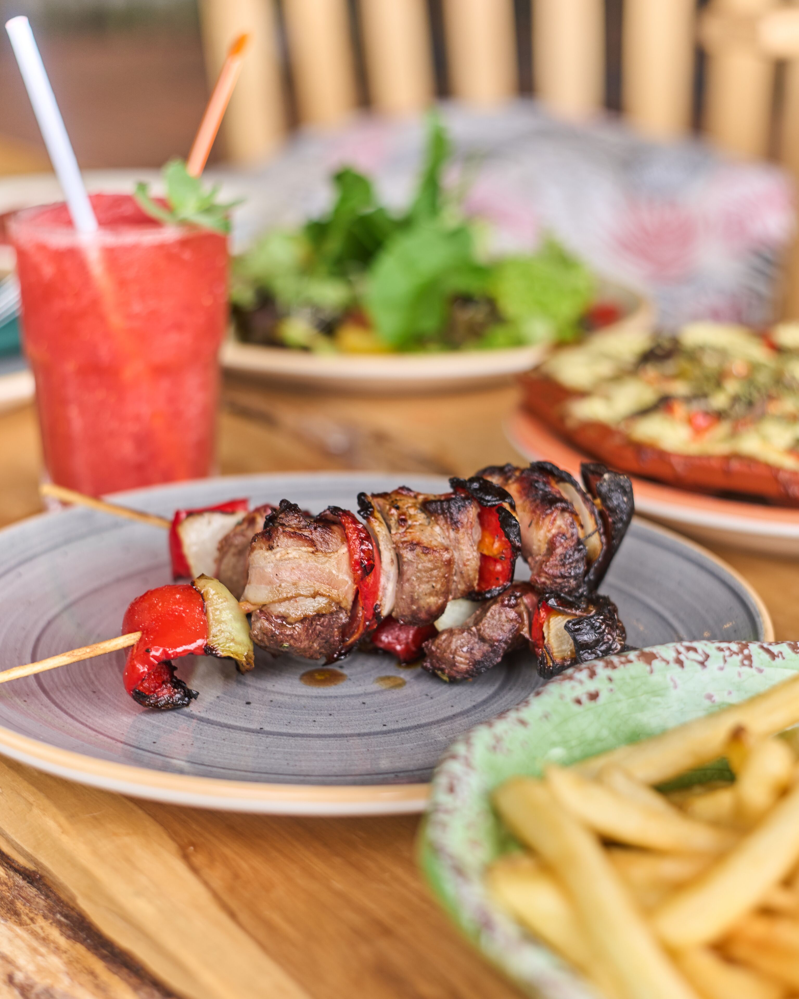 Ein Teller mit Spießen, Fleisch und Gemüse, ein Glas rotes Getränk, eine Schüssel Pommes und ein Salat.