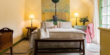 Un dormitorio con cama, lámparas de noche, un banco, cómoda, un cuadro y una ventana con cortinas.
