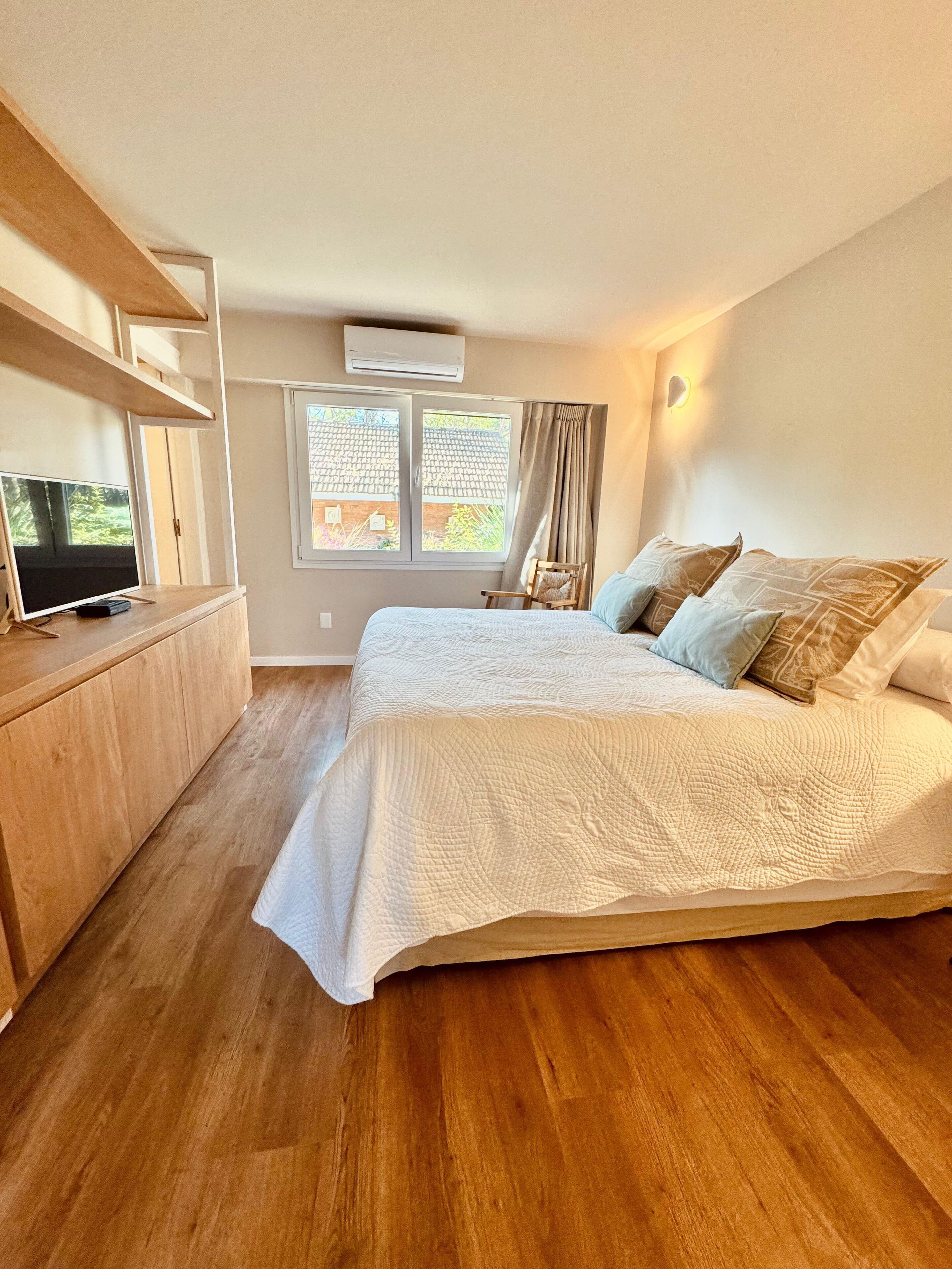 A bedroom with a large bed, a wooden dresser, a TV, and a window with curtains.