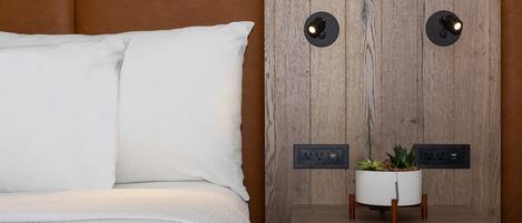 Una cama bien hecha con sábanas y almohadas blancas, una mesita de noche con una planta en maceta, y un enchufe en la pared.
