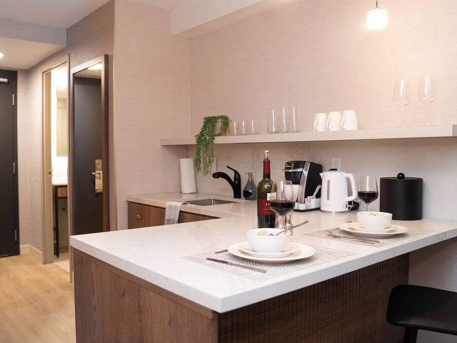 Une cuisine moderne avec un plan de travail blanc, une machine à café, une bouteille de vin et un saladier rempli de fruits.