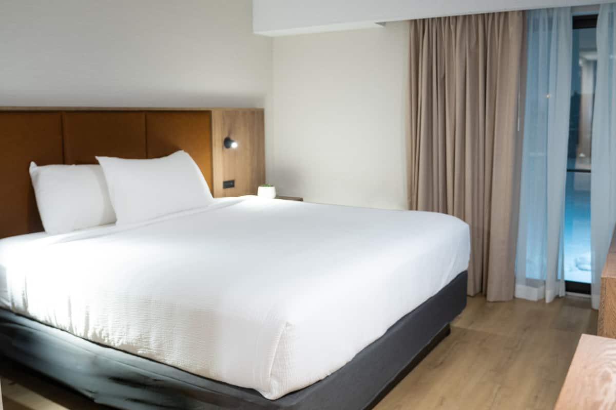 A modern hotel room with a large bed, wooden headboard, and a view of the outdoors through a window with curtains.