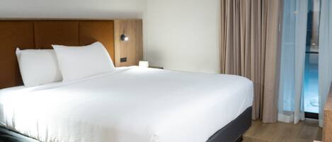 Une chambre d’hôtel moderne avec un grand lit, une tête de lit en bois et une vue sur l’extérieur par une fenêtre agrémentée de rideaux.
