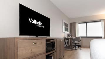 A hotel room with a flat-screen TV on a wooden cabinet, a desk with a chair, and a large window with curtains.