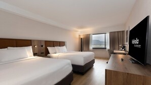 A hotel room with two beds, a desk, a flat-screen TV, and a view of the city.